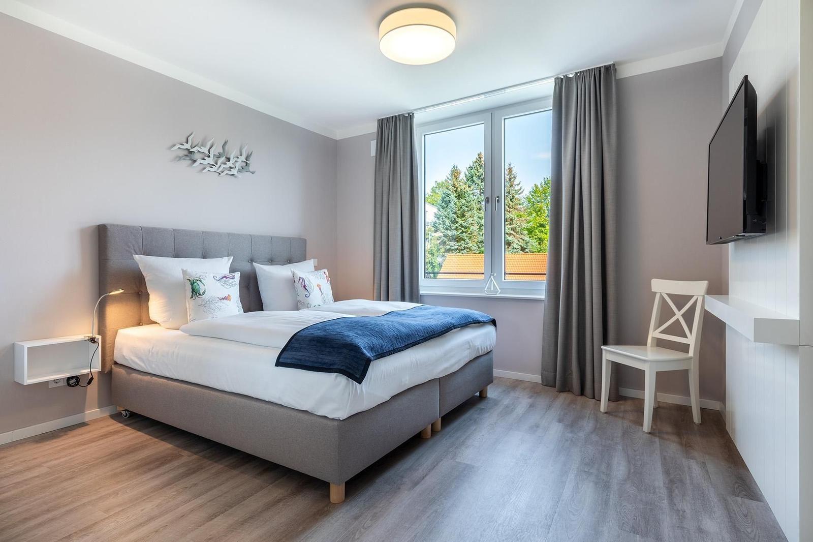 Schlafzimmer mit Doppelbett, Fenster, TV und Stuhl.