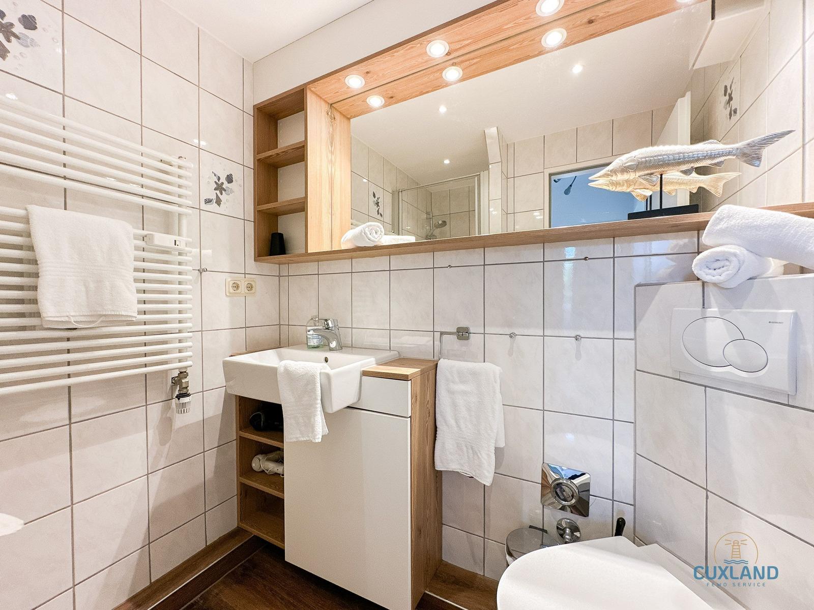Badezimmer mit Spiegel, Waschbecken und Toilette. Weiße Fliesen und Holzelemente.