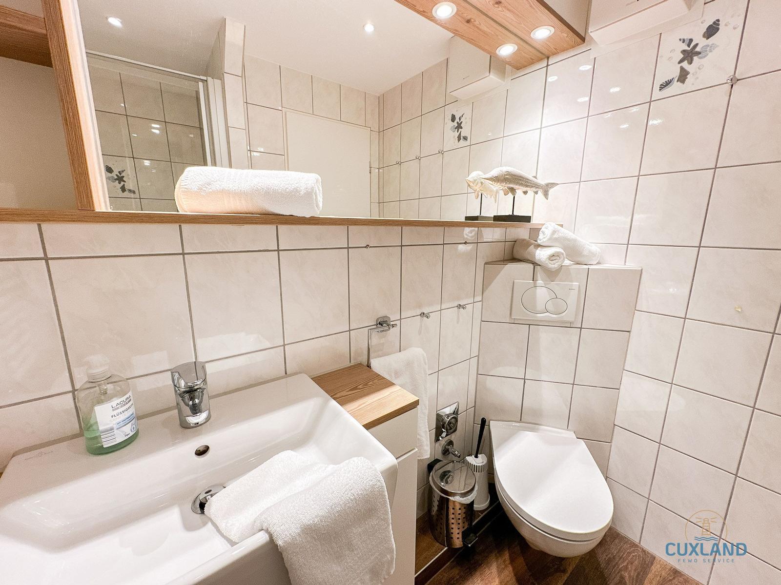 Badezimmer mit Waschbecken, Toilette und Dusche. Weiße Fliesen und Holzakzente.
