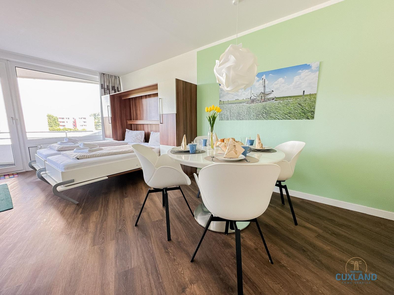 Ein Raum mit Esstisch, Stühlen und Bett. Wandbild und grüne Wand.