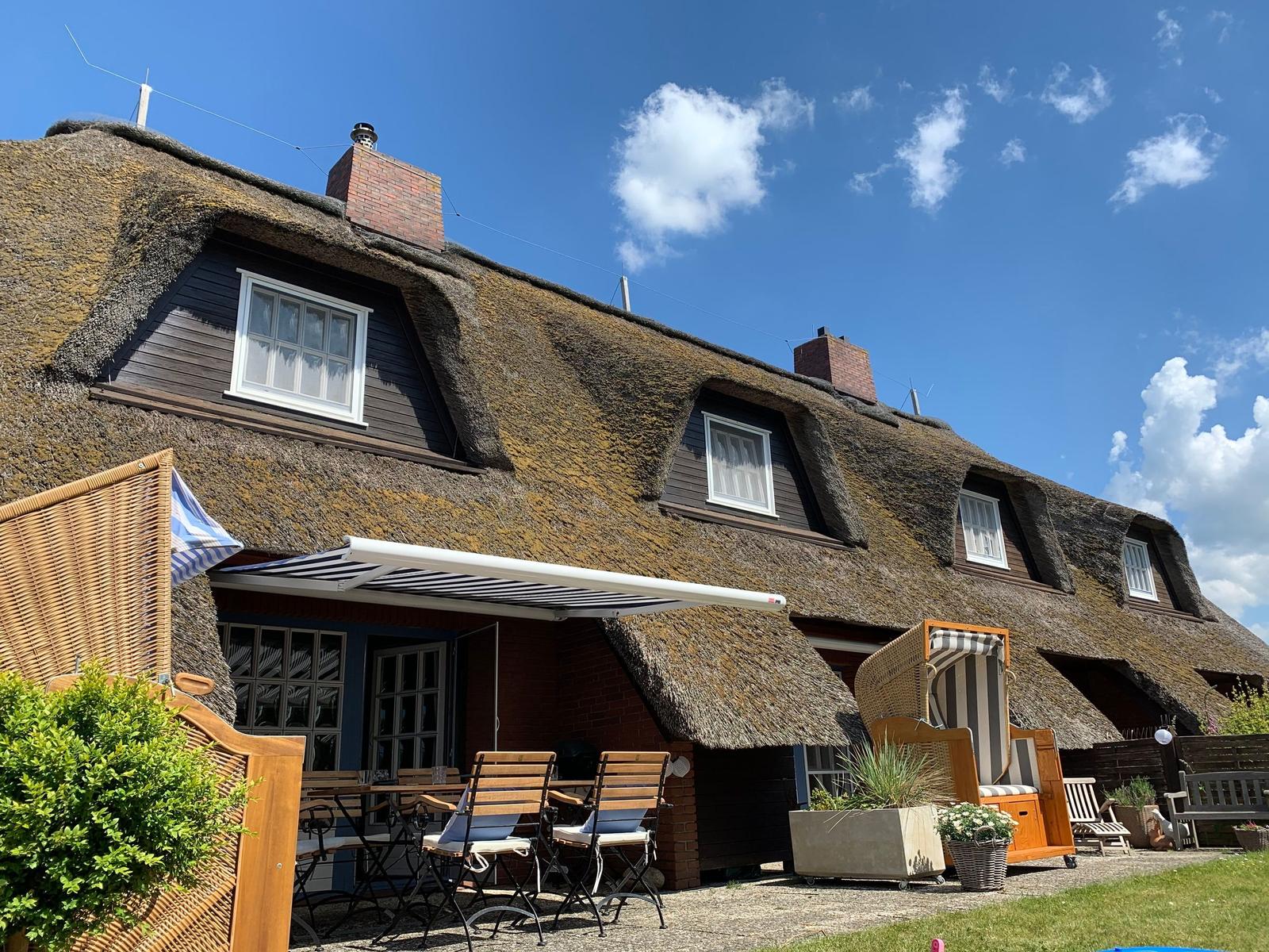 Dorfhaus mit Strohdach, Terrasse und Gartenmöbel unter blauem Himmel.