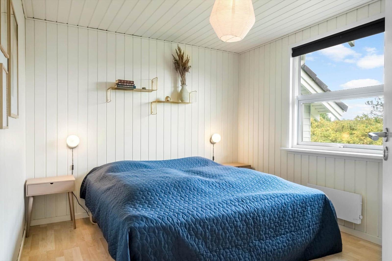 Schlafzimmer mit blauer Bettdecke, Holzboden und Fenster mit Blick auf Grün.