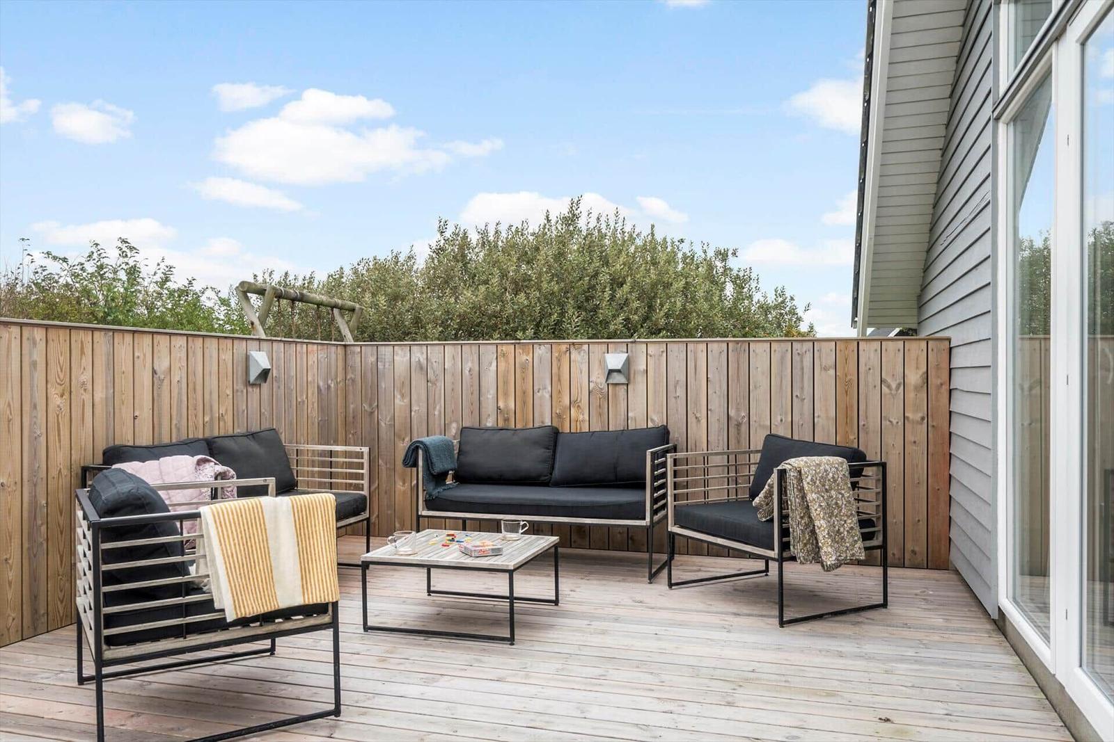 Terrasse mit Sitzgruppe, Tisch und Holzgeländer. Hintergrund: Bäume und blauer Himmel.