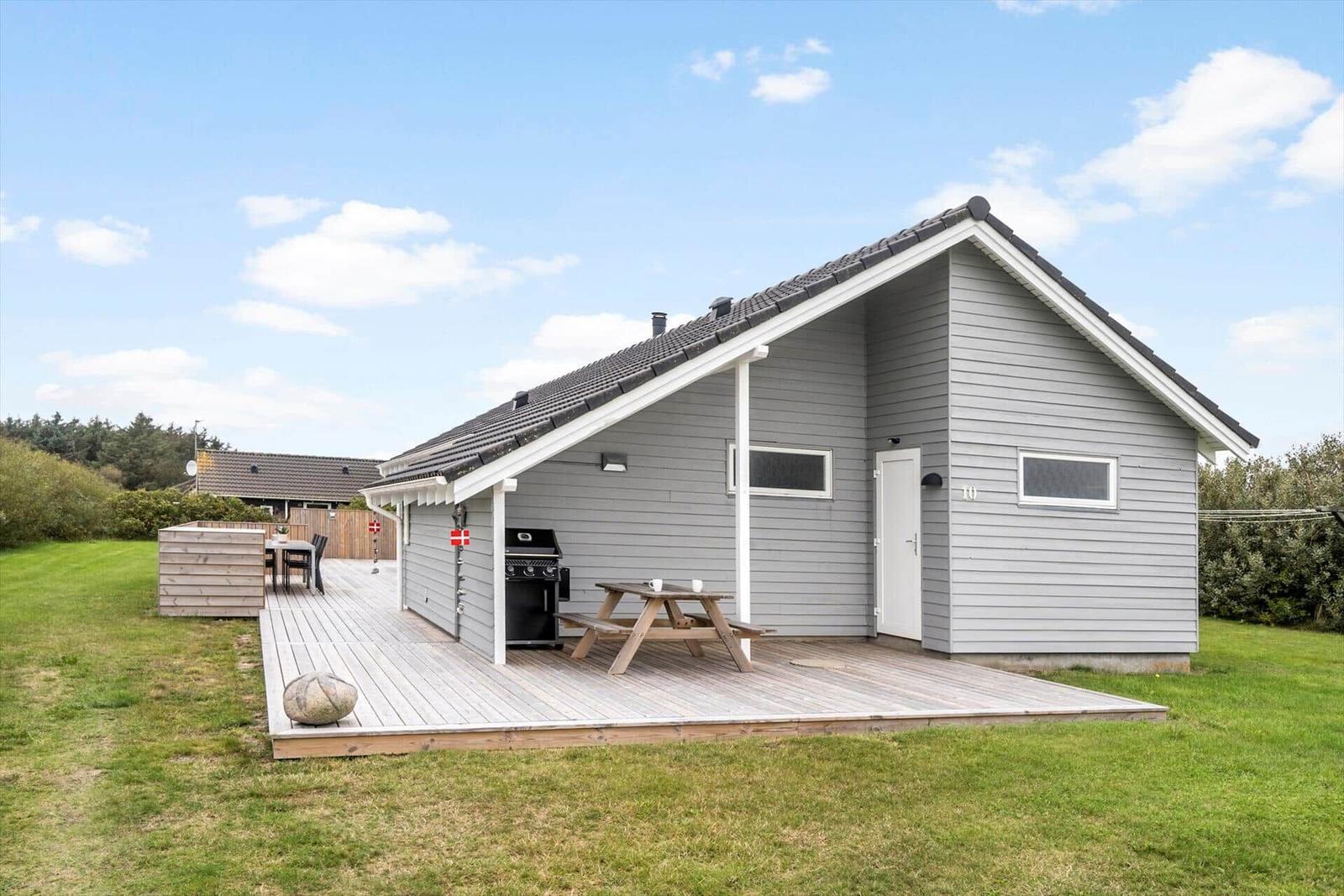 Ein graues Ferienhaus mit Terrasse, Grill und Picknicktisch. Grün umgeben.