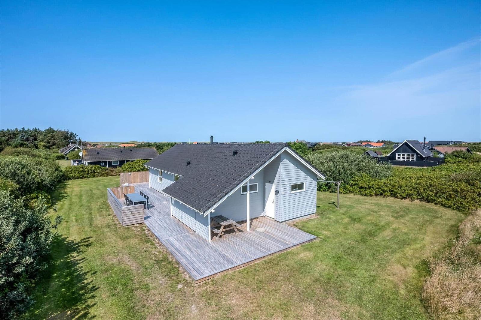 Ein hellblaues Haus mit Holzterrasse und Garten. Hintergrund: Grünflächen und andere Häuser.