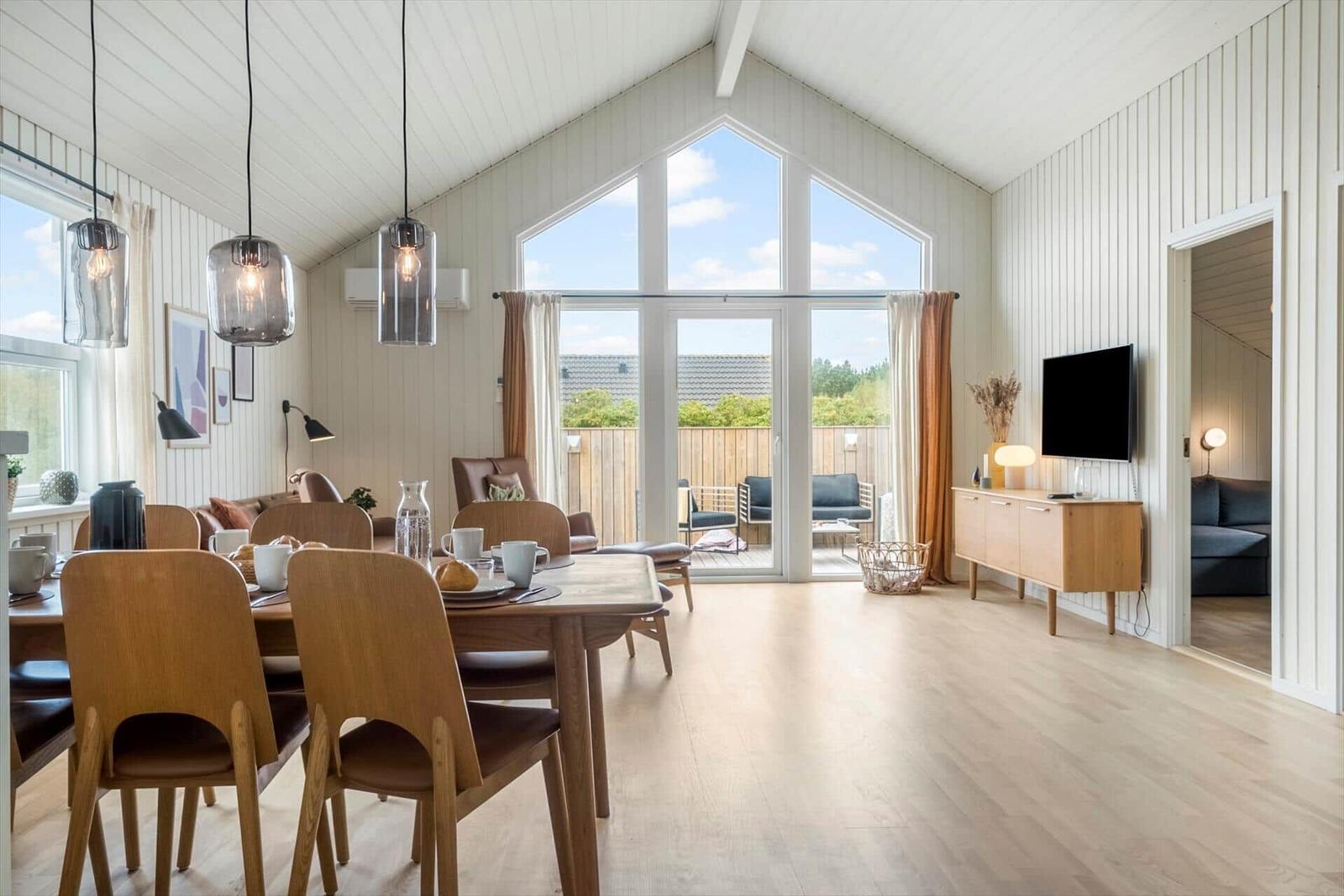 Wohn- und Essbereich mit Holzboden, großen Fenstern und TV. Terrasse mit Sitzmöbeln.