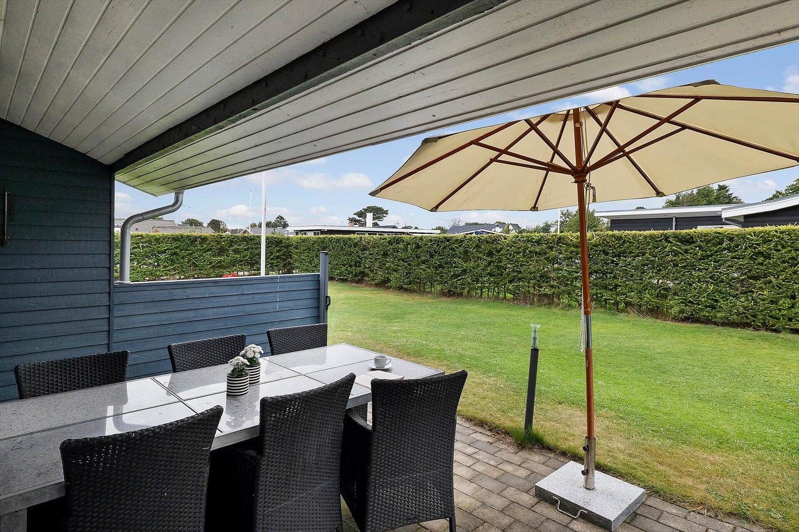 Terrasse mit Tisch, Stühlen und Sonnenschirm. Grünfläche und Hecke im Hintergrund.