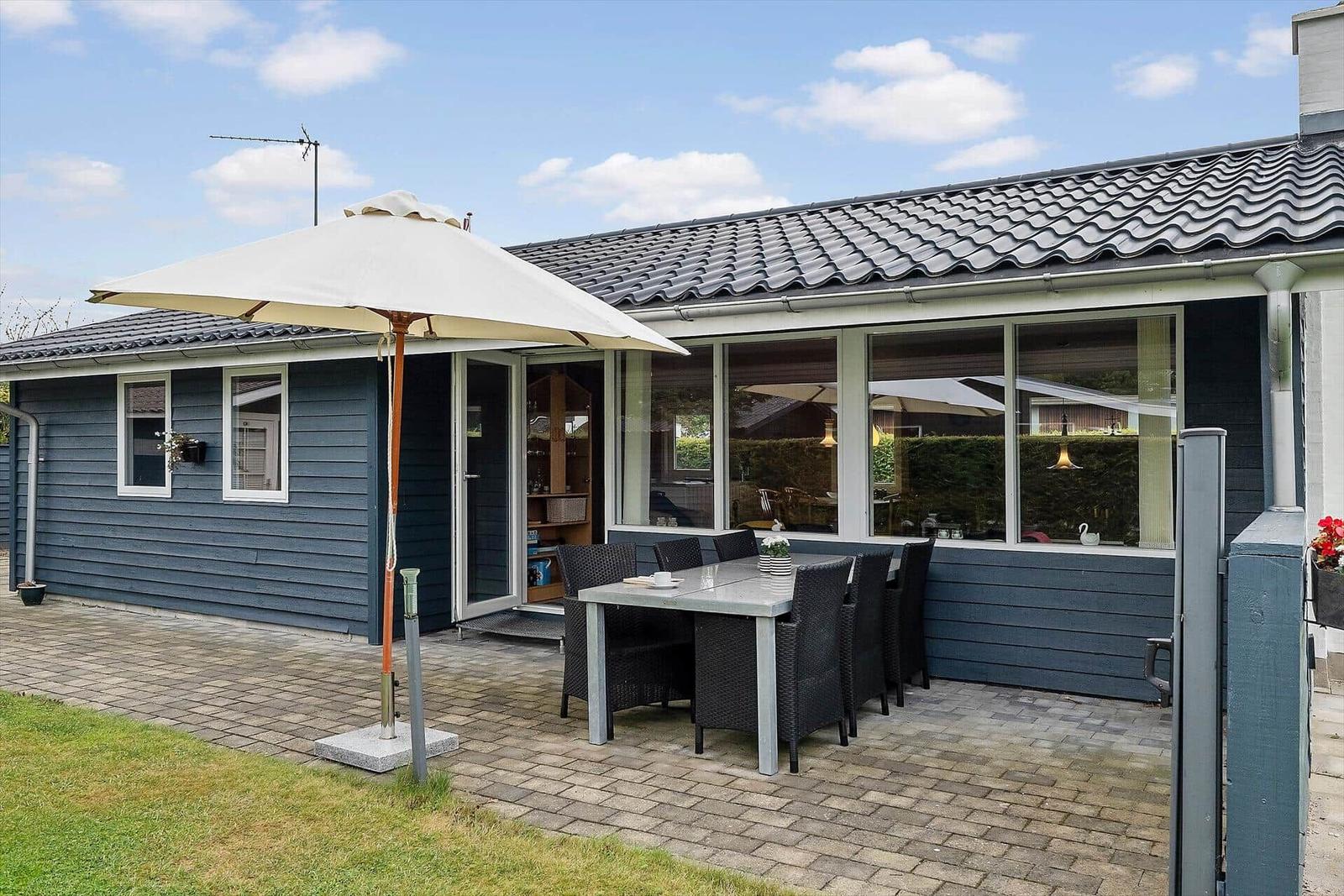Terrasse mit Tisch, Stühlen und Sonnenschirm vor einem Haus mit großen Fenstern.