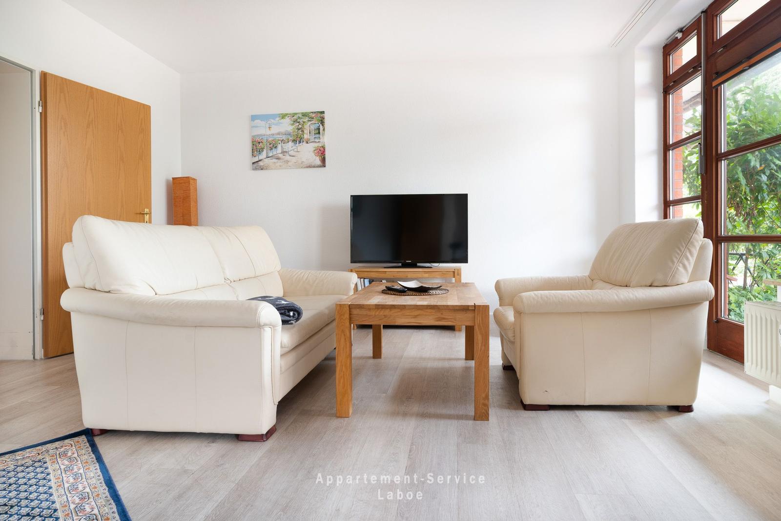 Wohnzimmer mit Sofa, Sessel und Fernseher. Holztisch und große Fenster.