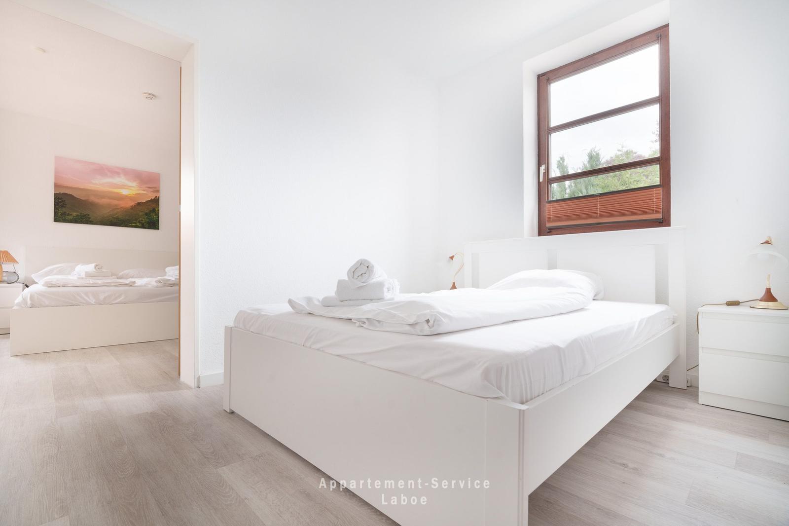 Zimmer mit weißem Bett, Fenster und Nachttisch. Durchgang zu weiterem Schlafzimmer.