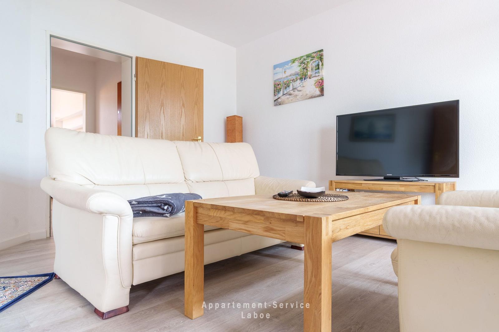 Wohnzimmer mit Sofa, Holztisch und Fernseher. Wandbild und Holztür sichtbar.