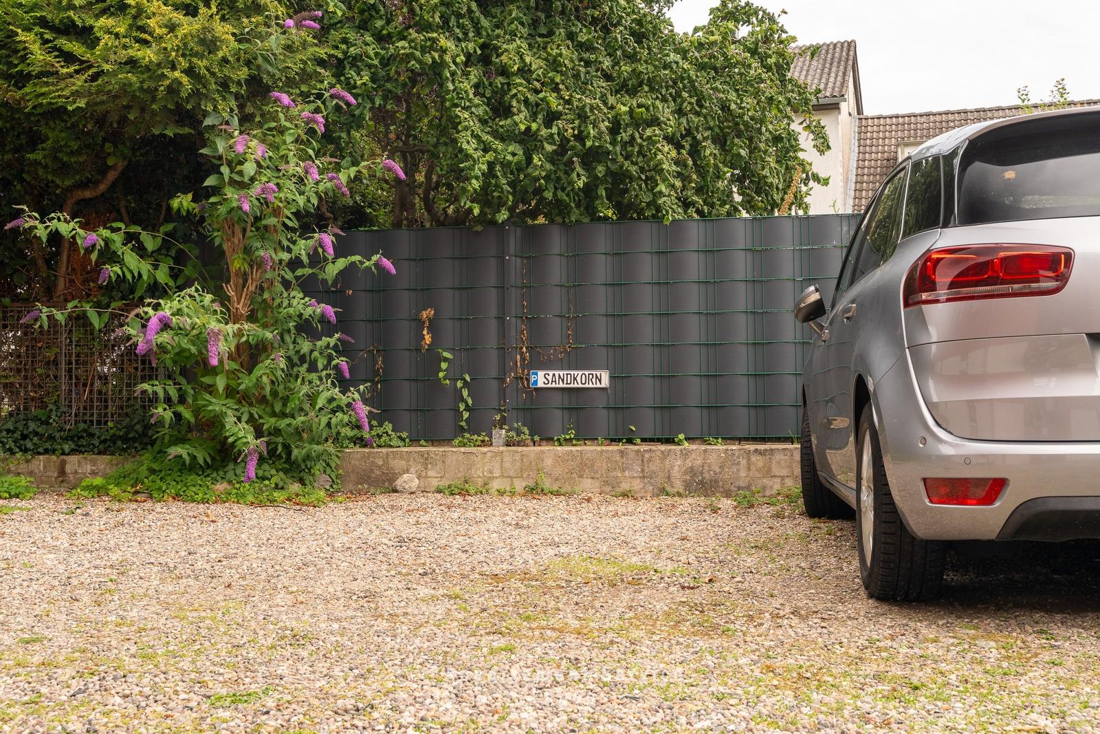 Ein silberner Wagen parkt auf Kies vor einem Zaun mit einer Pflanze und einem Schild.