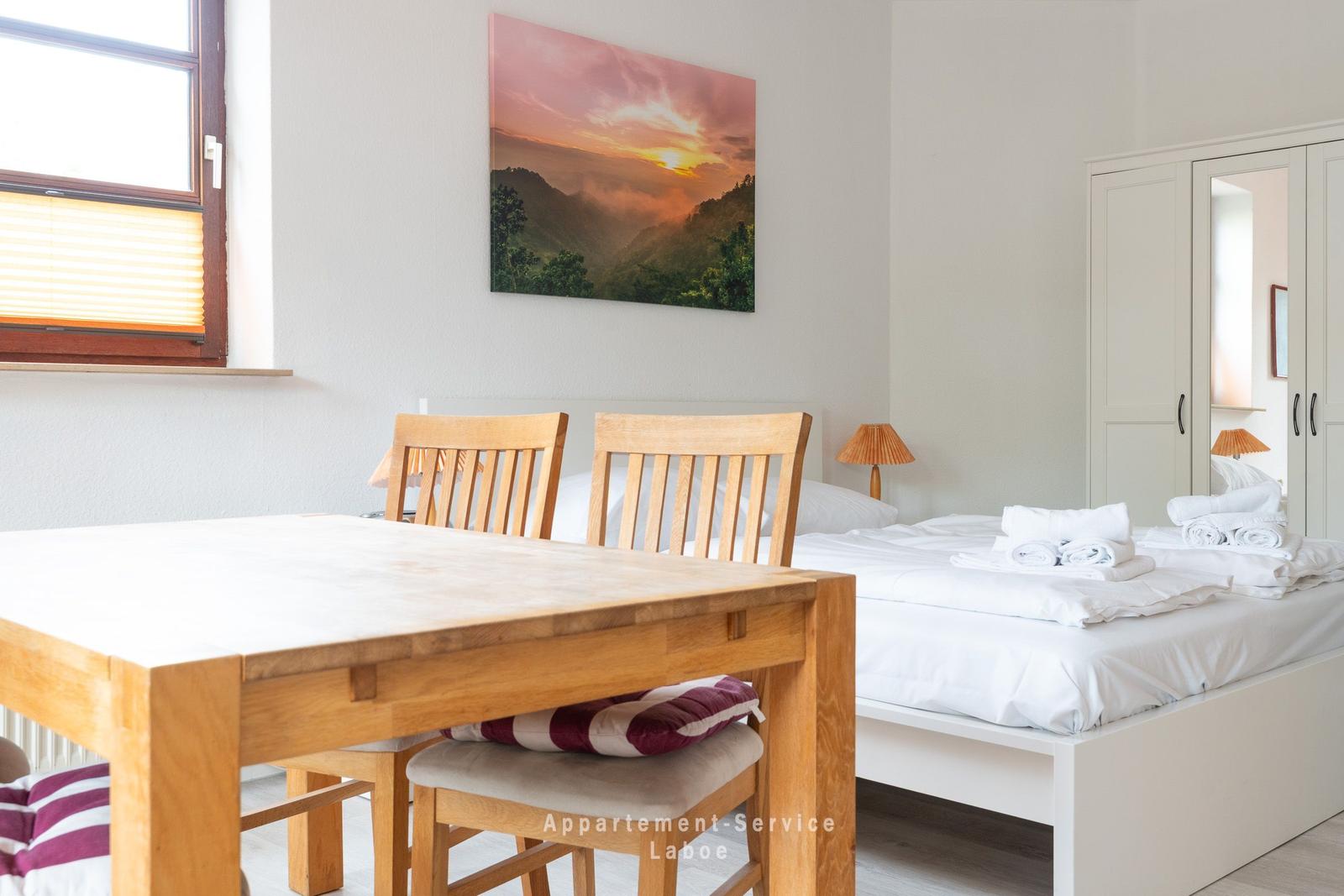 Schlafzimmer mit Bett, Tisch und Stühlen. Wandbild zeigt Sonnenuntergang.