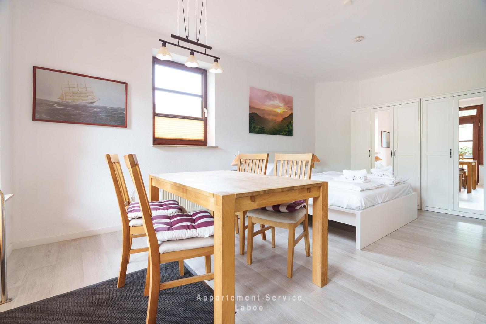 Ein Raum mit Tisch, Stühlen, Bett und Schränken. An den Wänden hängen Bilder.