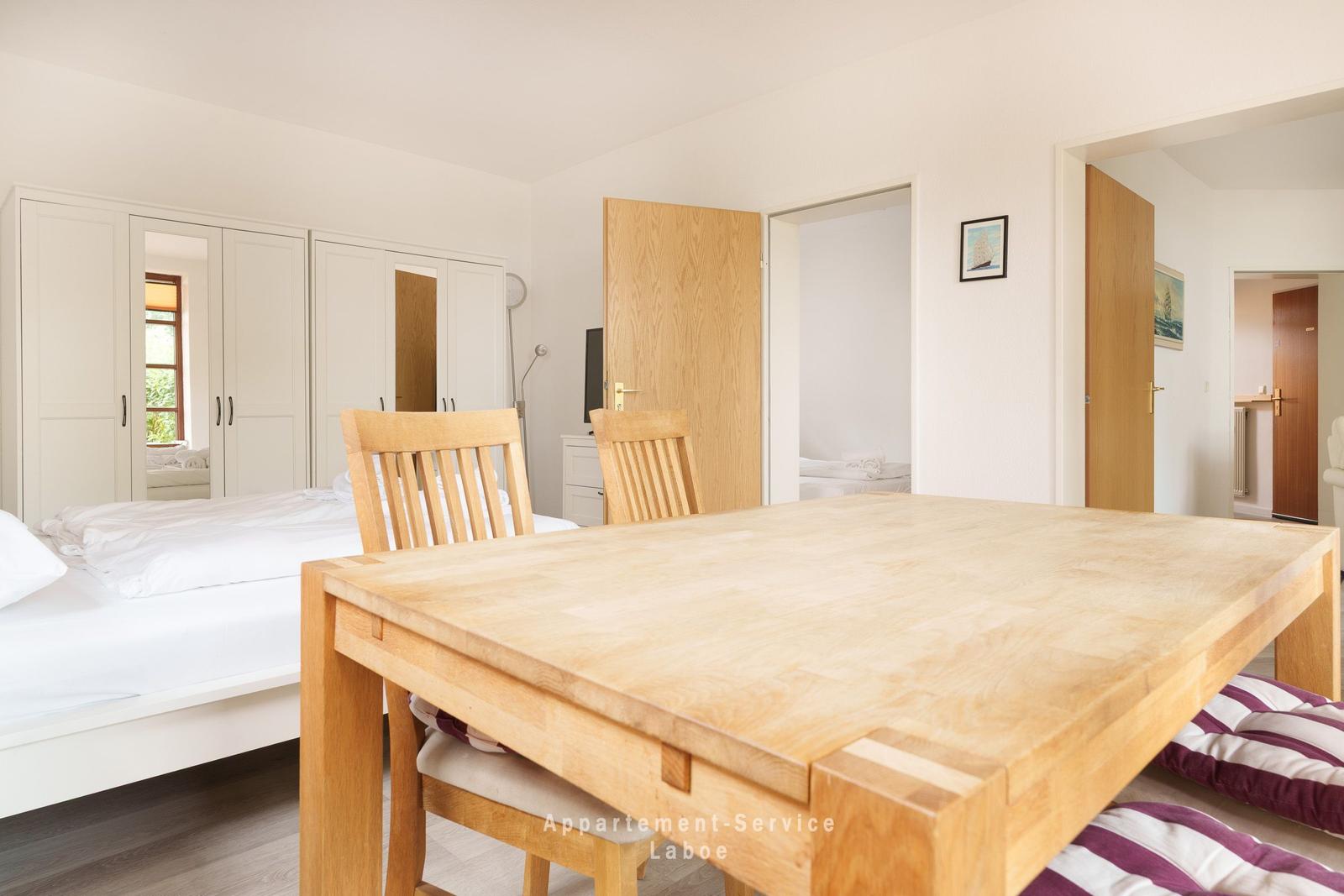 Holztisch mit Stühlen vor Bett und Kleiderschrank. Türen führen zu weiteren Räumen.