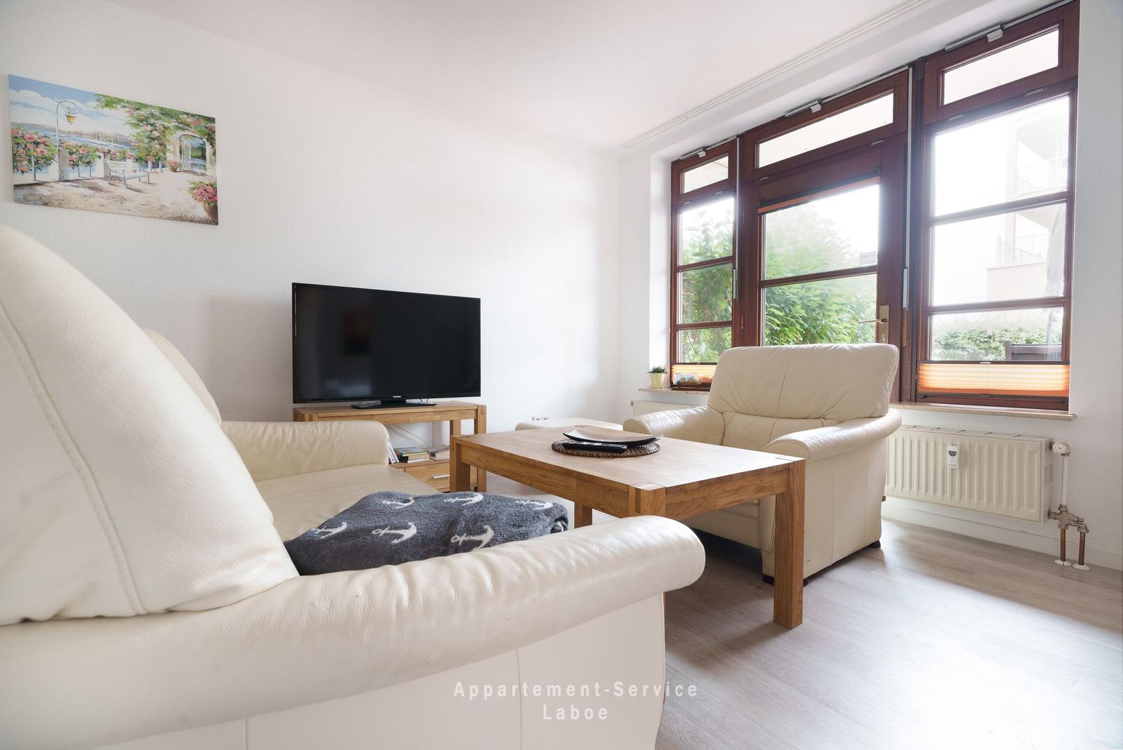 Wohnzimmer mit Sofa, Sessel, Fernseher und großen Fenstern.