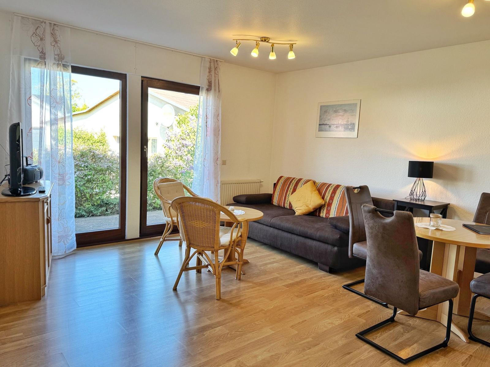 Wohnzimmer mit Sofa, Esstisch und Terrassentür. Holzboden und Fensterfront.