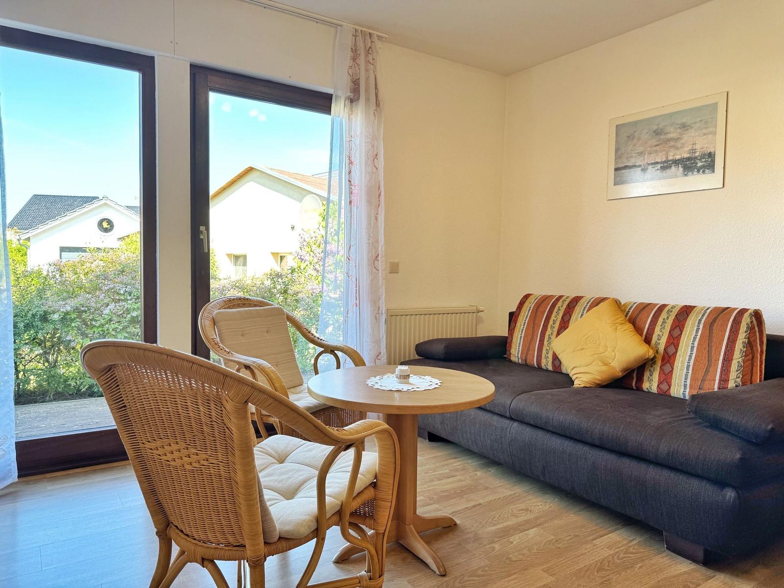 Wohnzimmer mit Sofa, Tisch und Stühlen. Durchgang zur Terrasse mit Blick auf Garten.