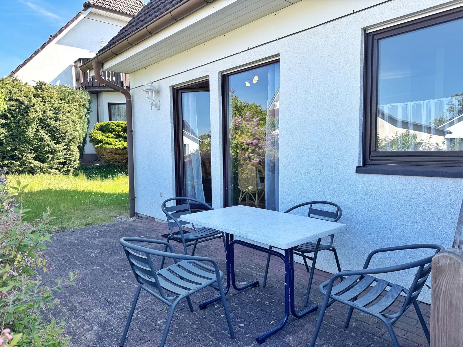 Terrasse mit Tisch und vier Stühlen vor weißem Haus mit Fenstern.