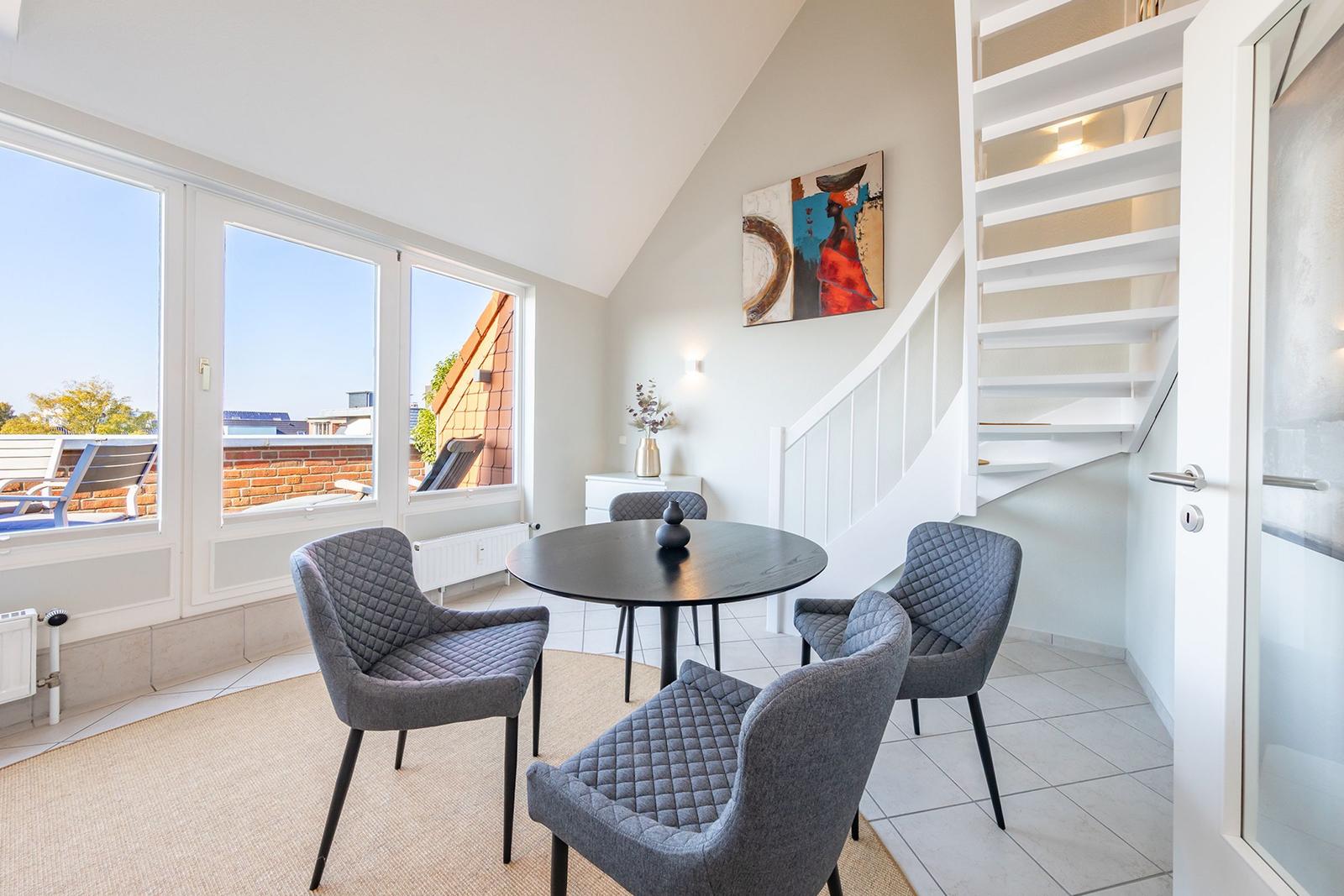 Essbereich mit runden Tisch und vier Stühlen. Weiße Treppe und große Fenster zur Terrasse.