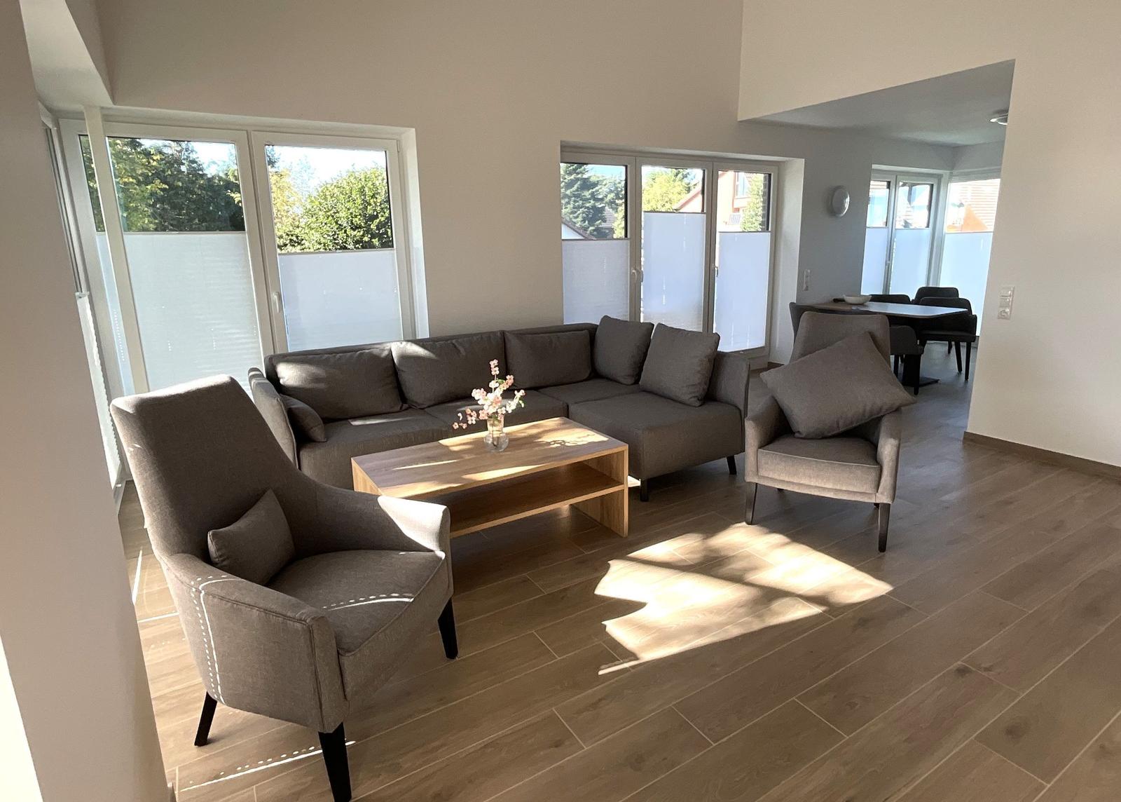 Wohnzimmer mit Sofa, zwei Sesseln und Holztisch. Durchgänge zu Essbereich und Terrasse.