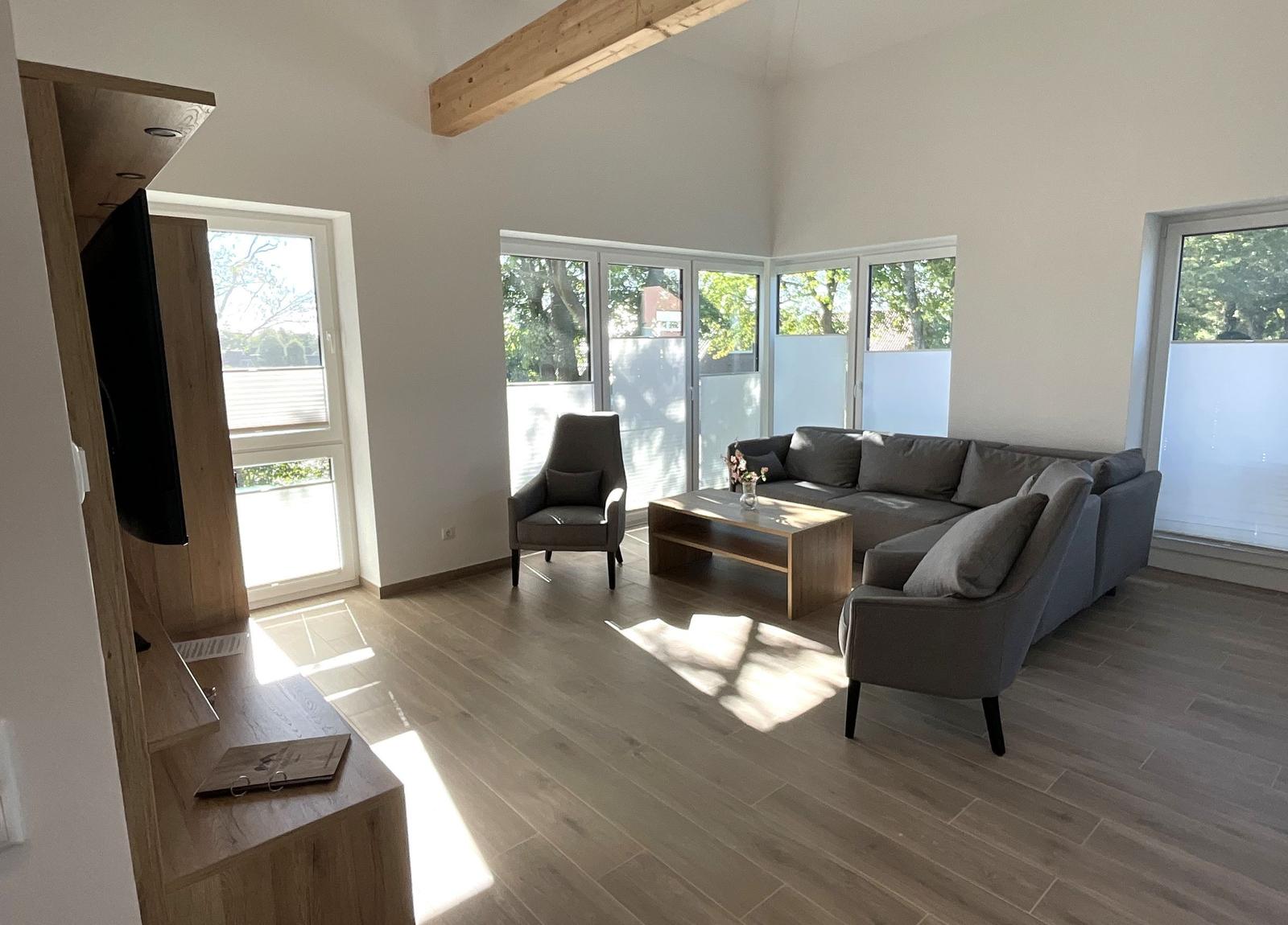 Wohnzimmer mit Sofa, Sessel und Holzboden. Große Fenster und Holzbalken.