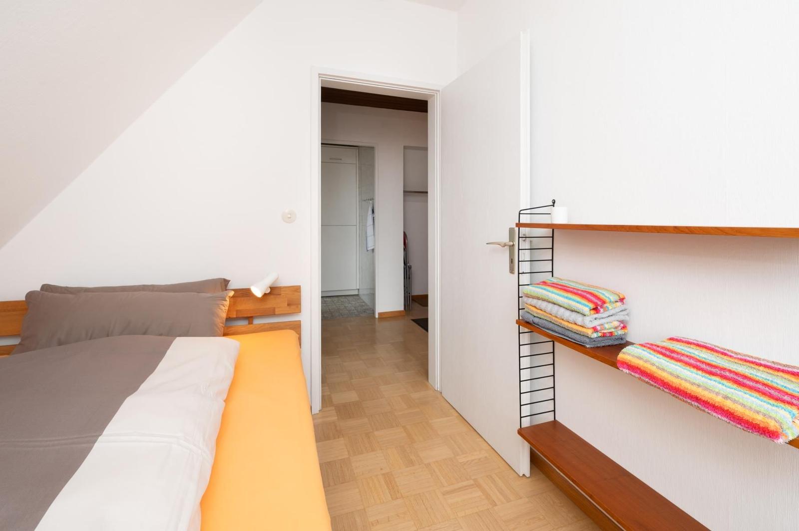 Schlafzimmer mit Bett, Holzregal und Bad im Hintergrund.