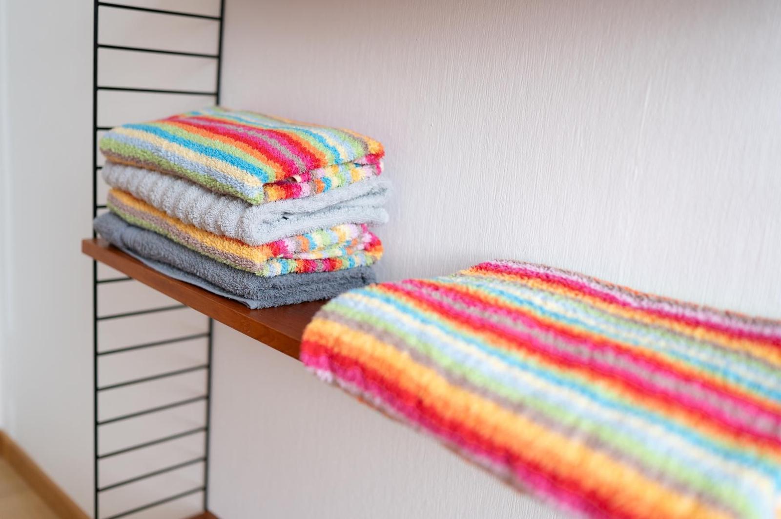 Farbige Handtücher liegen auf einem Holzregal. Ein weiteres Handtuch liegt daneben.