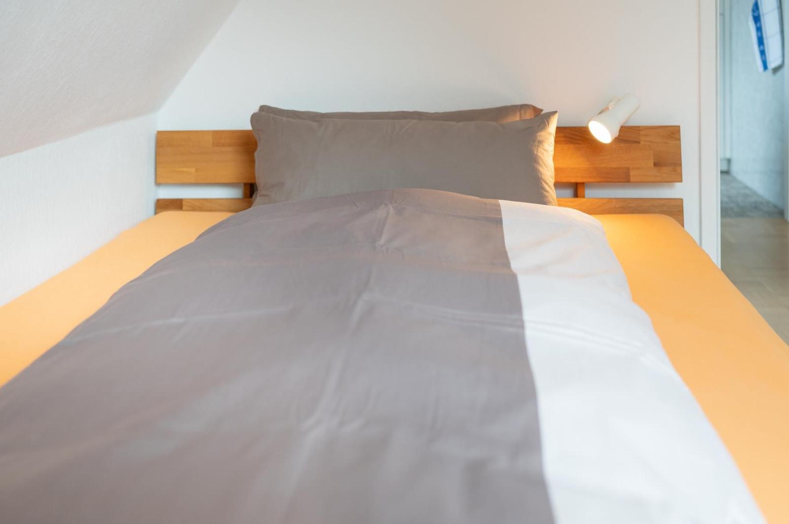 Ein Bett mit Holzkopfteil, grauer Bettwäsche und einer Wandlampe.