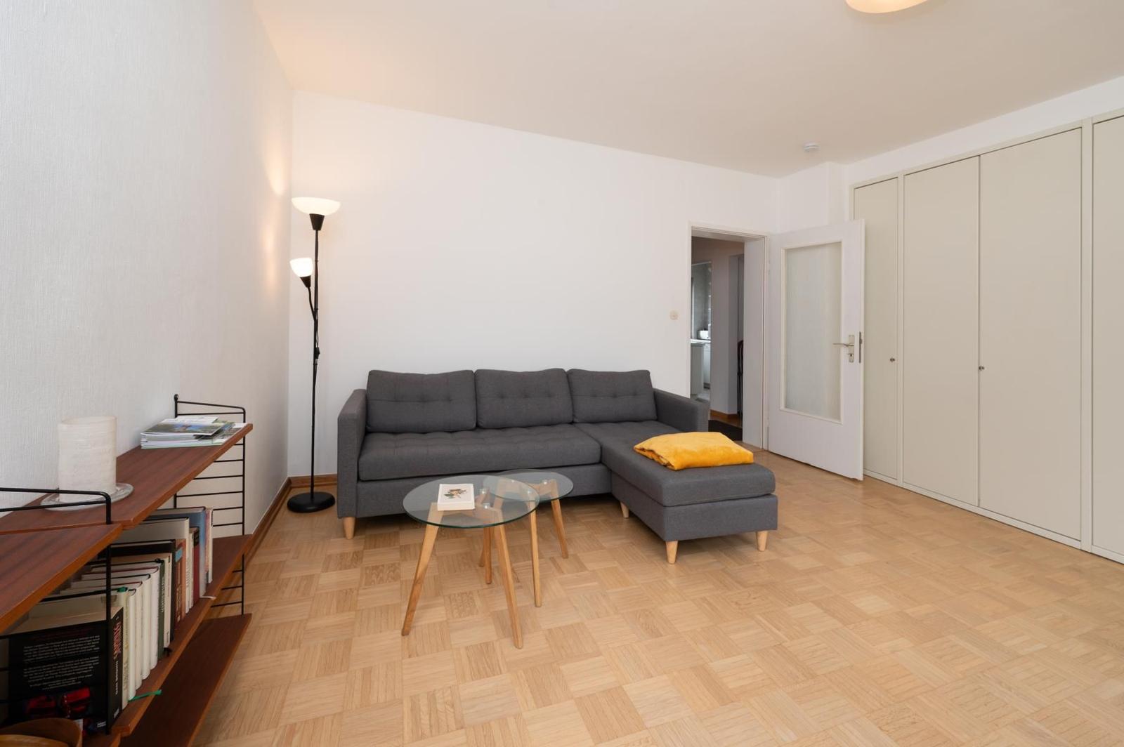 Wohnzimmer mit grauem Sofa, Glas-Tisch und Holzboden. An der Wand steht ein Bücherregal.