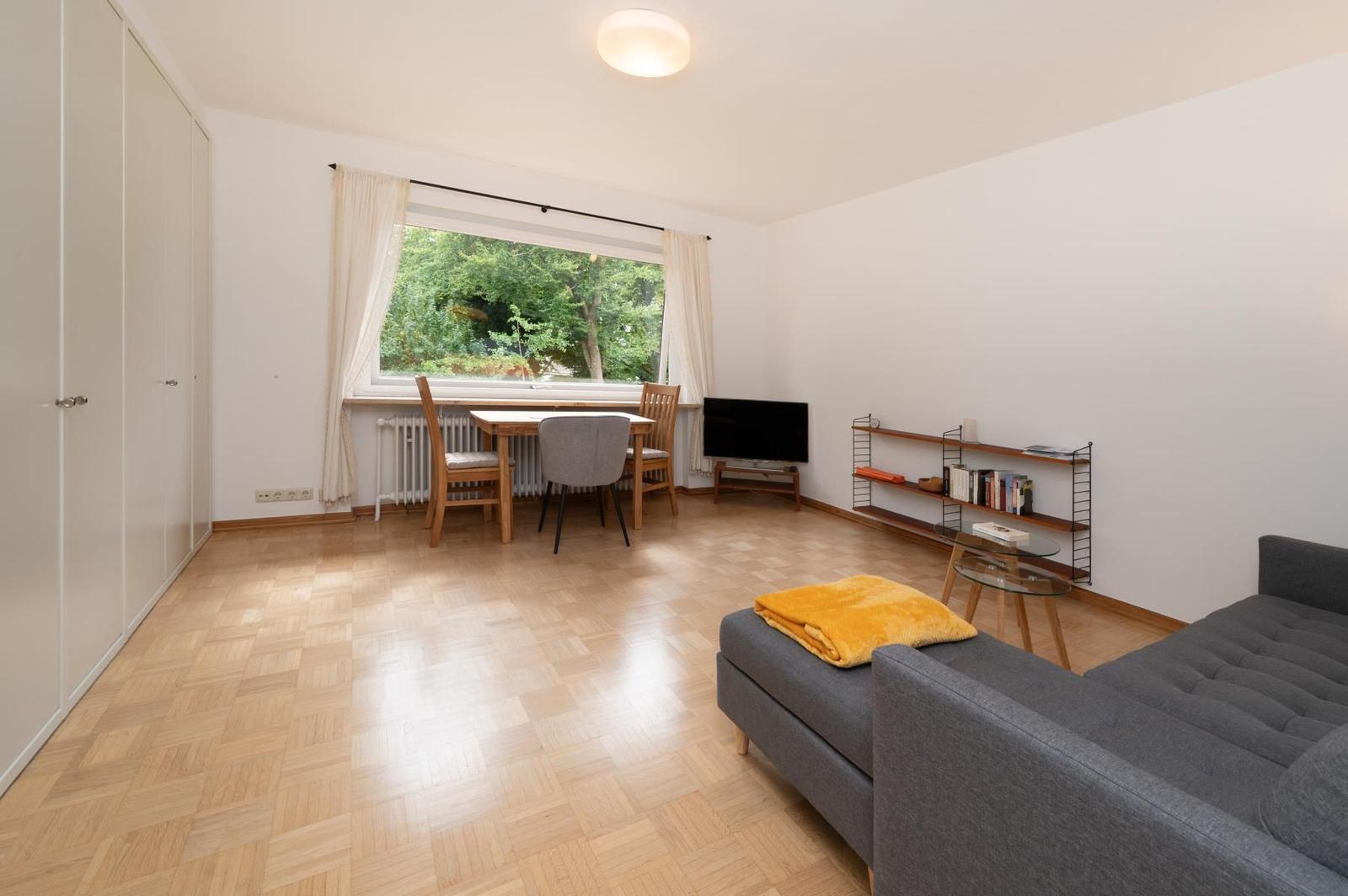 Wohnzimmer mit Sofa, Esstisch und TV. Holzboden und Fenster mit Blick auf Grün.