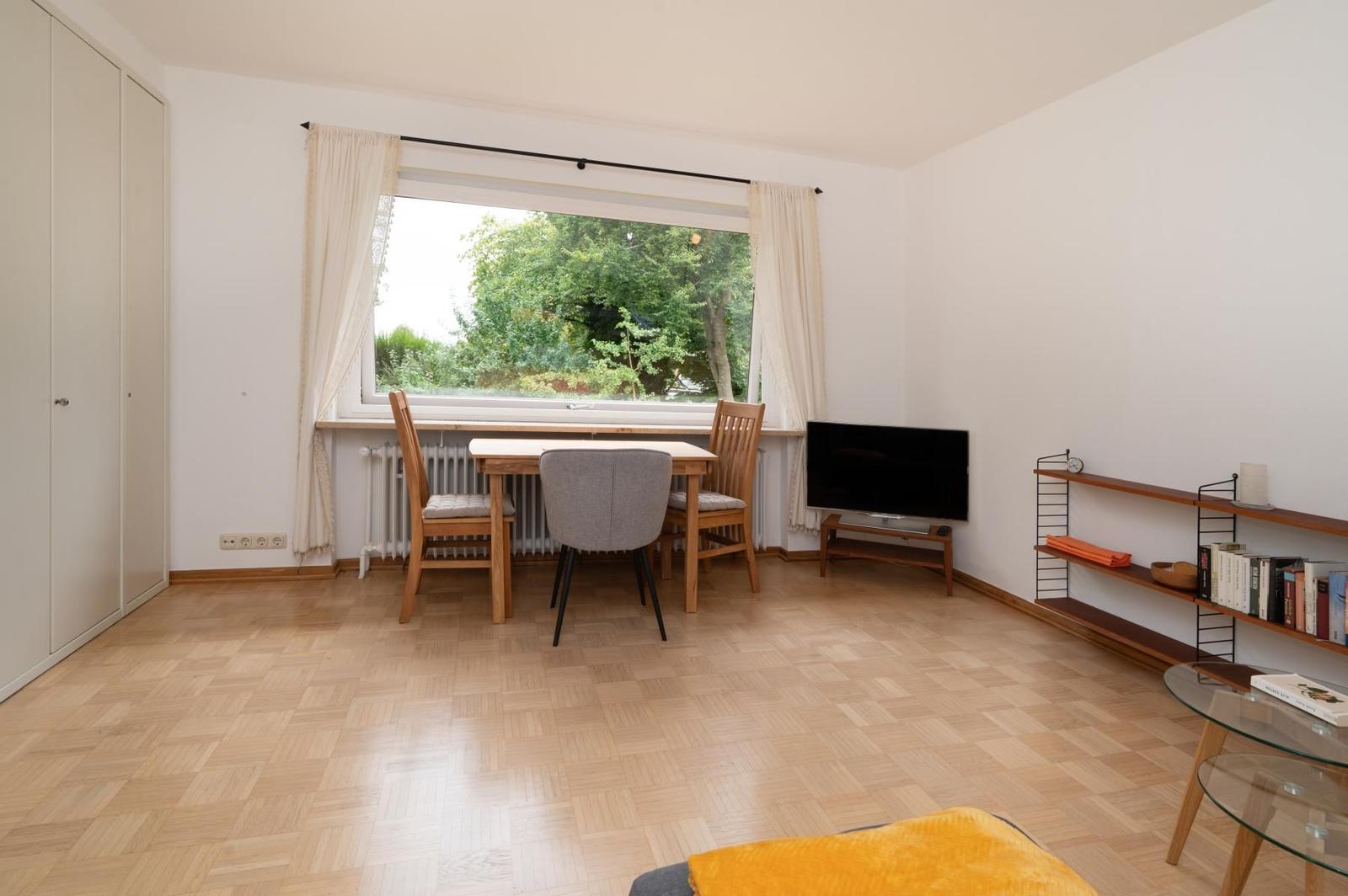 Raum mit Esstisch, TV und Bücherregal. Holzboden und Fenster mit Blick auf Grün.