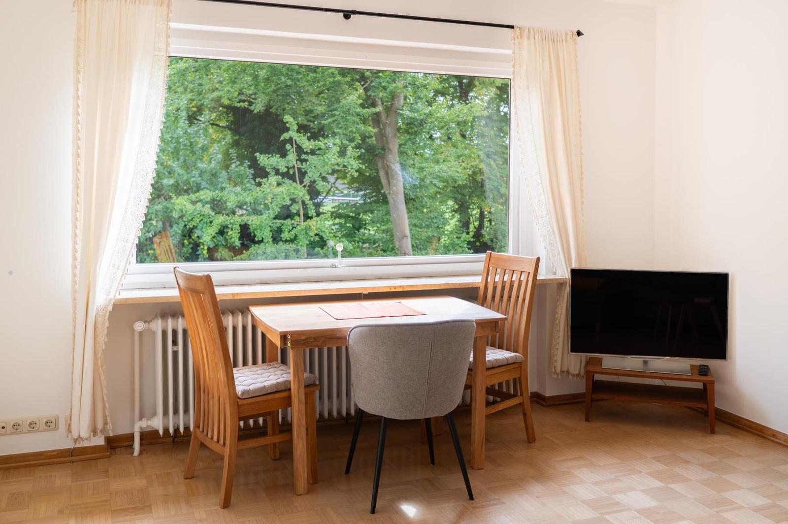 Esszimmer mit Tisch, Stühlen und TV. Große Fenster mit Blick auf Bäume.