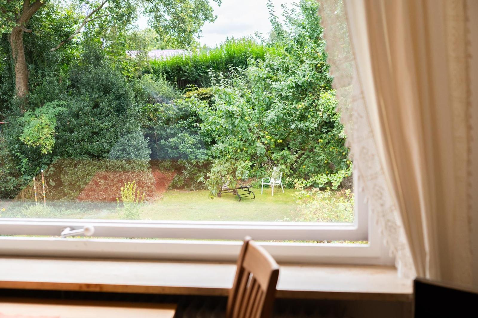 Durch das Fenster blickt man auf einen grünen Garten mit Bäumen und einem weißen Stuhl.