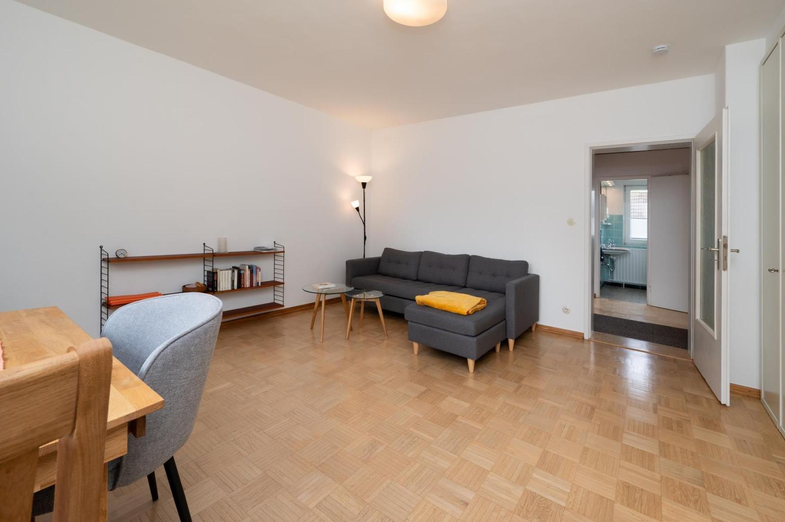 Wohnzimmer mit Sofa, Tisch und Bücherregal. Holzfußboden und Tür zum Bad.