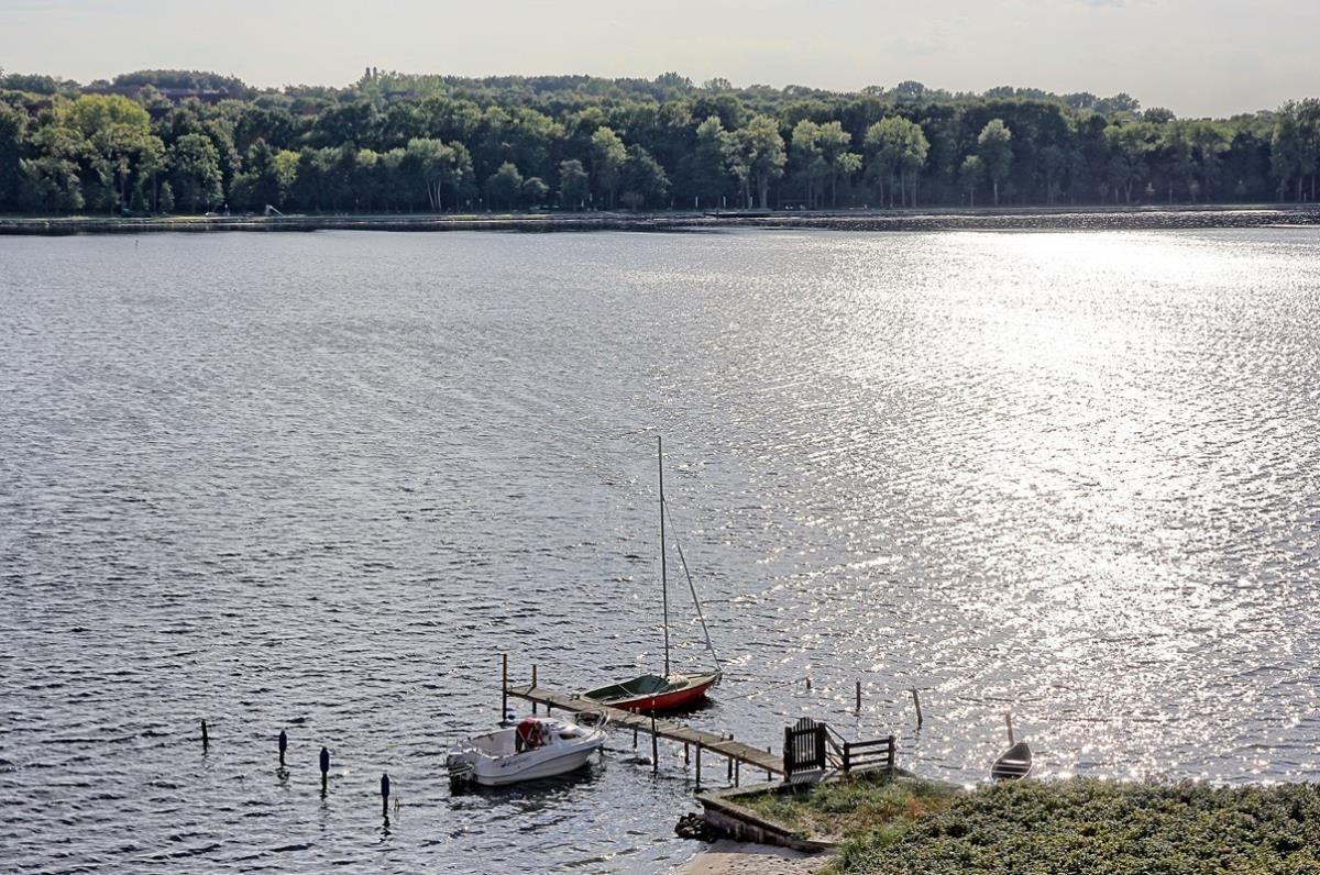 Bootshafen mit zwei Booten an Steg und grüner Uferlinie im Hintergrund.