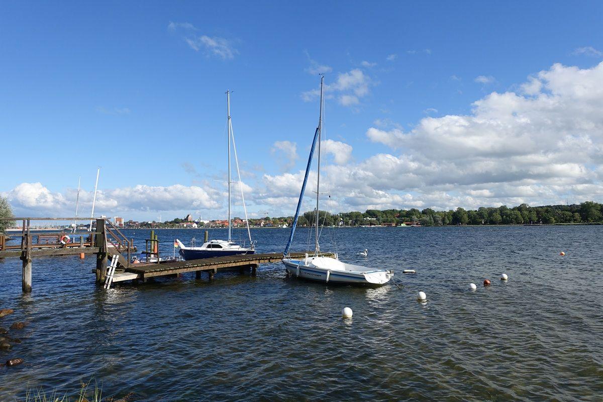 Zwei Segelboote liegen an einem Holzsteg im Wasser. Im Hintergrund sind Bäume und ein klarer Himmel.
