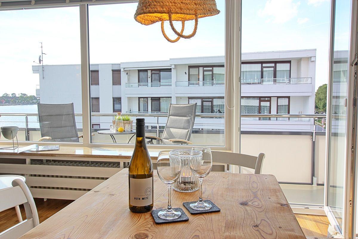 Holztisch mit Weinglas und Flasche vor großem Fenster mit Blick auf Balkon und Wasser.