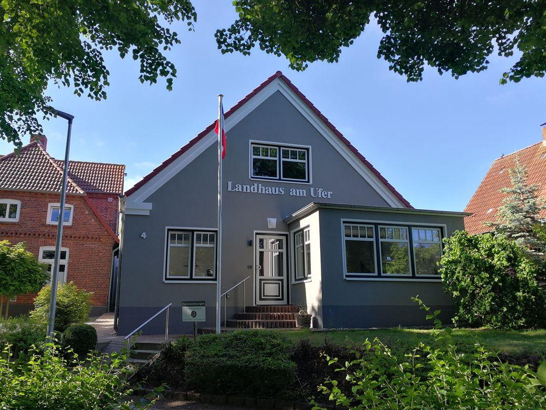 Landhaus am Ufer mit grauer Fassade und rotem Dach unter blauem Himmel.