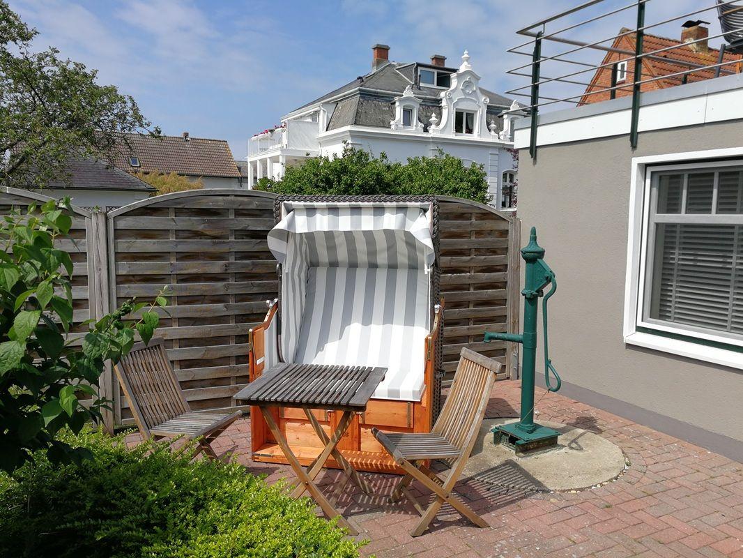 Terrasse mit Strandkorb, Holzstühlen und Brunnen. Hintergrund: Holzzaun und Gebäude.