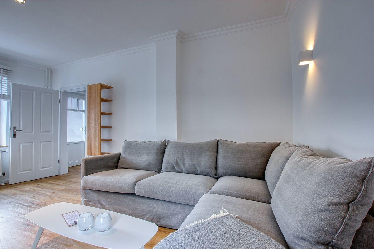 Wohnzimmer mit grauem Sofa, weißen Wänden und Holzboden. Es gibt eine Couchtisch und eine Wandlampe.