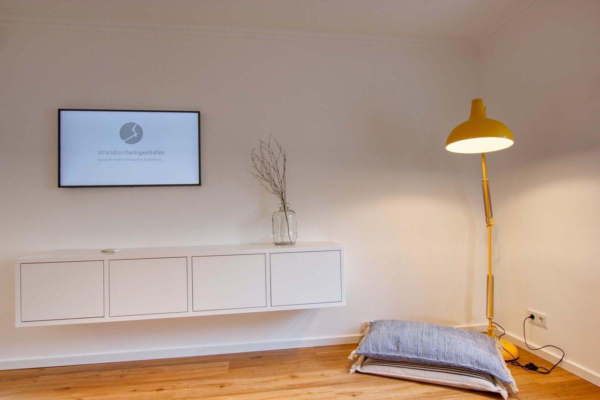 Wohnzimmer mit Wand-TV, weißen Schränken und gelber Stehlampe.