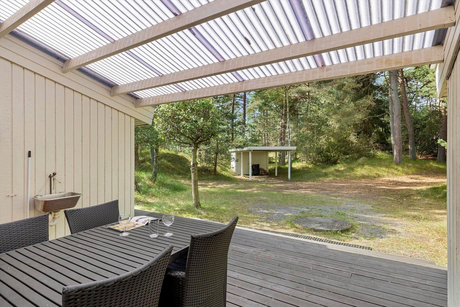 Terrasse mit Tisch und Stühlen unter Dach. Blick in den Wald mit kleinem Holzhaus.