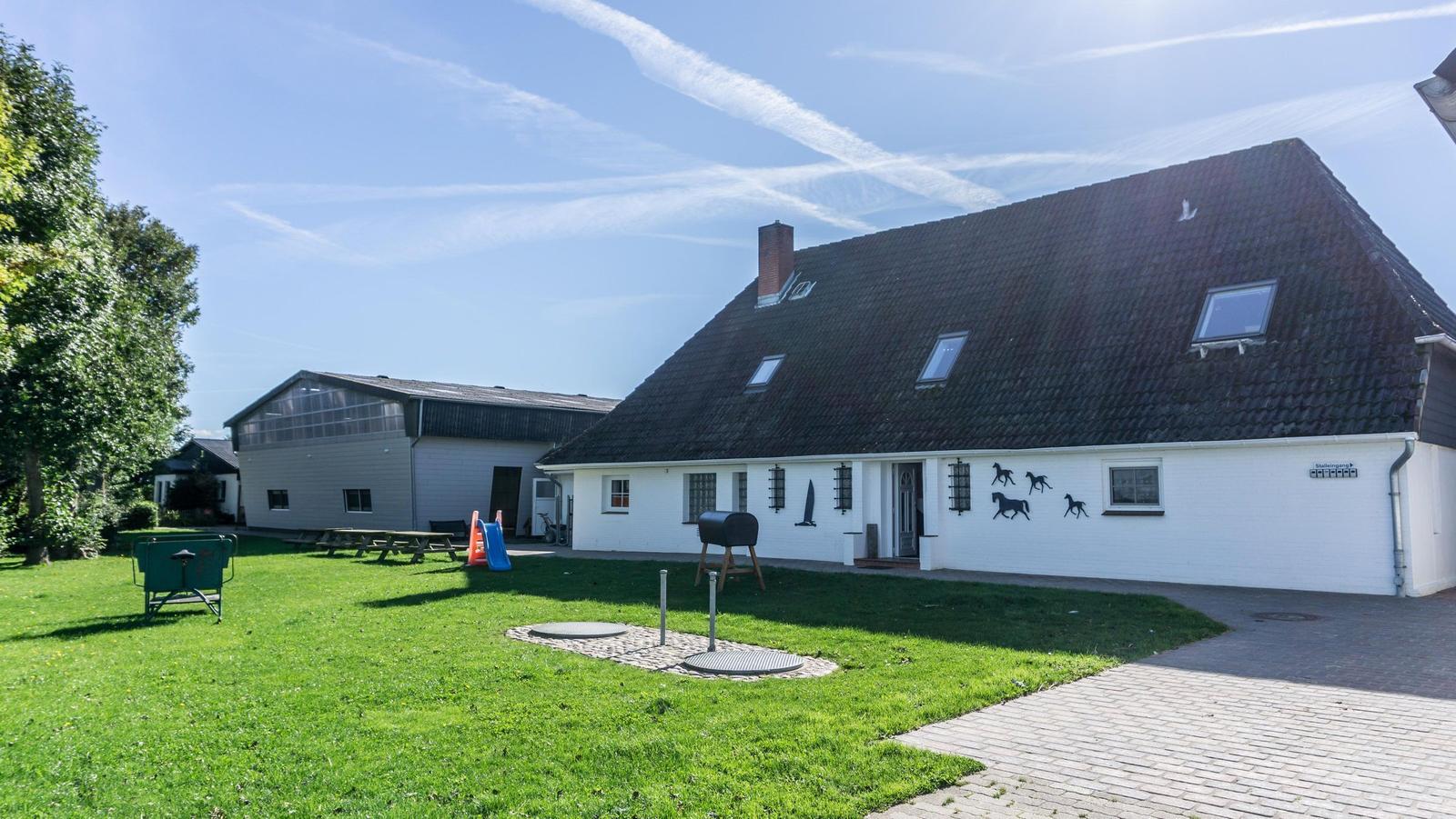 Haus mit weißer Fassade und braunem Dach. Grünfläche mit Spielplatz und Grill.