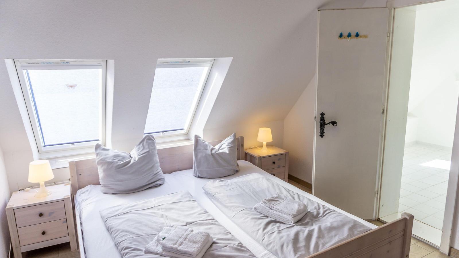 Schlafzimmer mit Dachfenstern, Bett, Nachttischen und Lampen. Tür führt in Bad.
