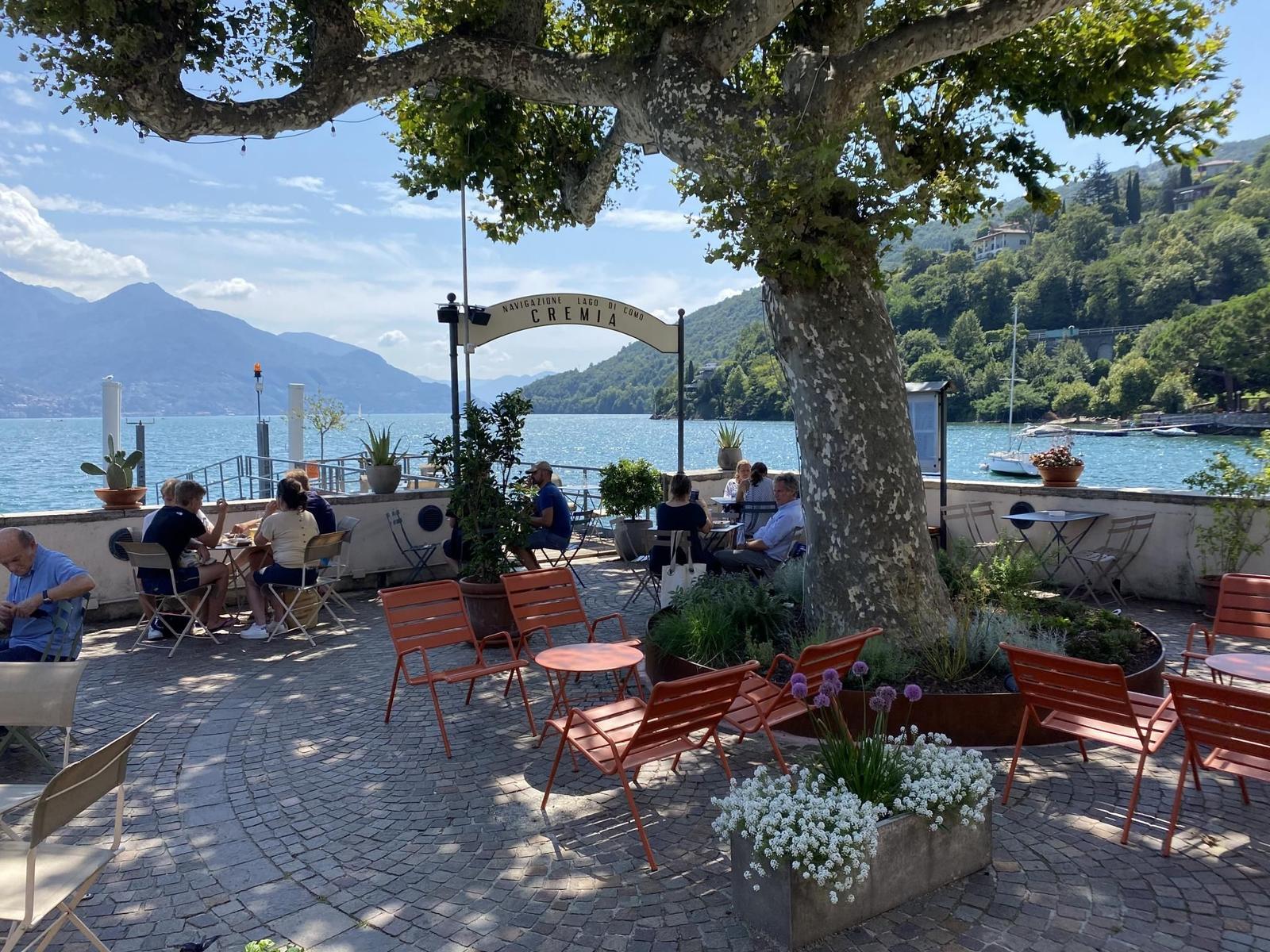 Terrace with tables and chairs by the lake. Large tree and view of water.
