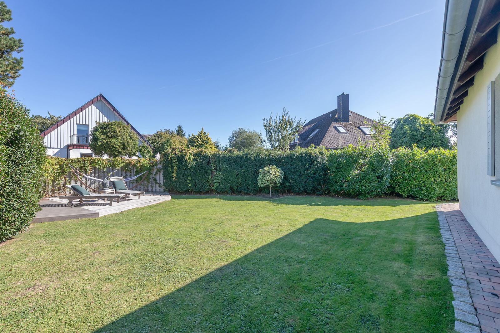 Hinterhof mit Rasenfläche, Holzterrasse und Liegen. Umzäunt mit Hecke. Himmel klar blau.