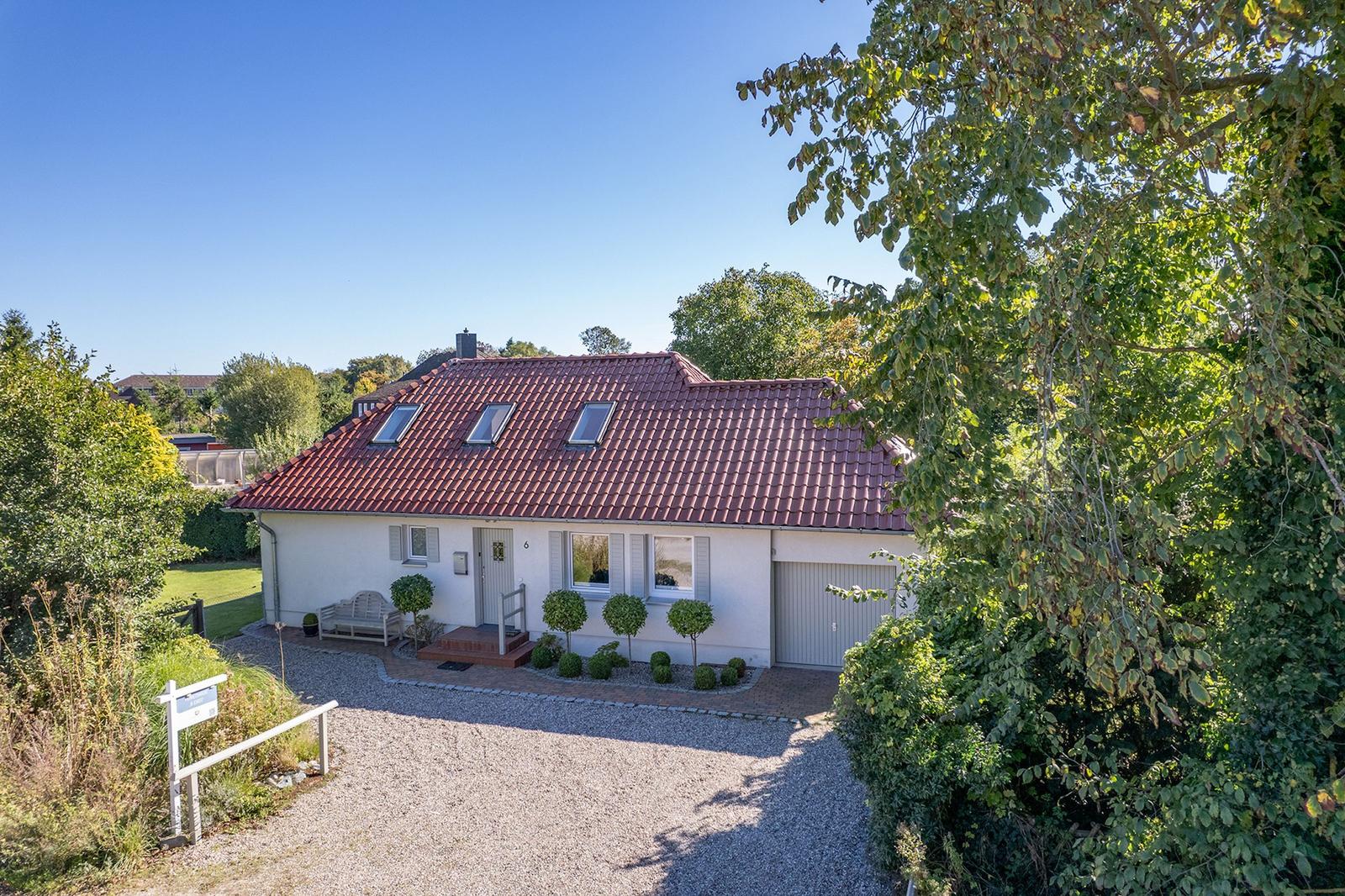 Ein weißes Haus mit rotem Dach und Garagenbereich. Umgeben von Grün und Bäumen.