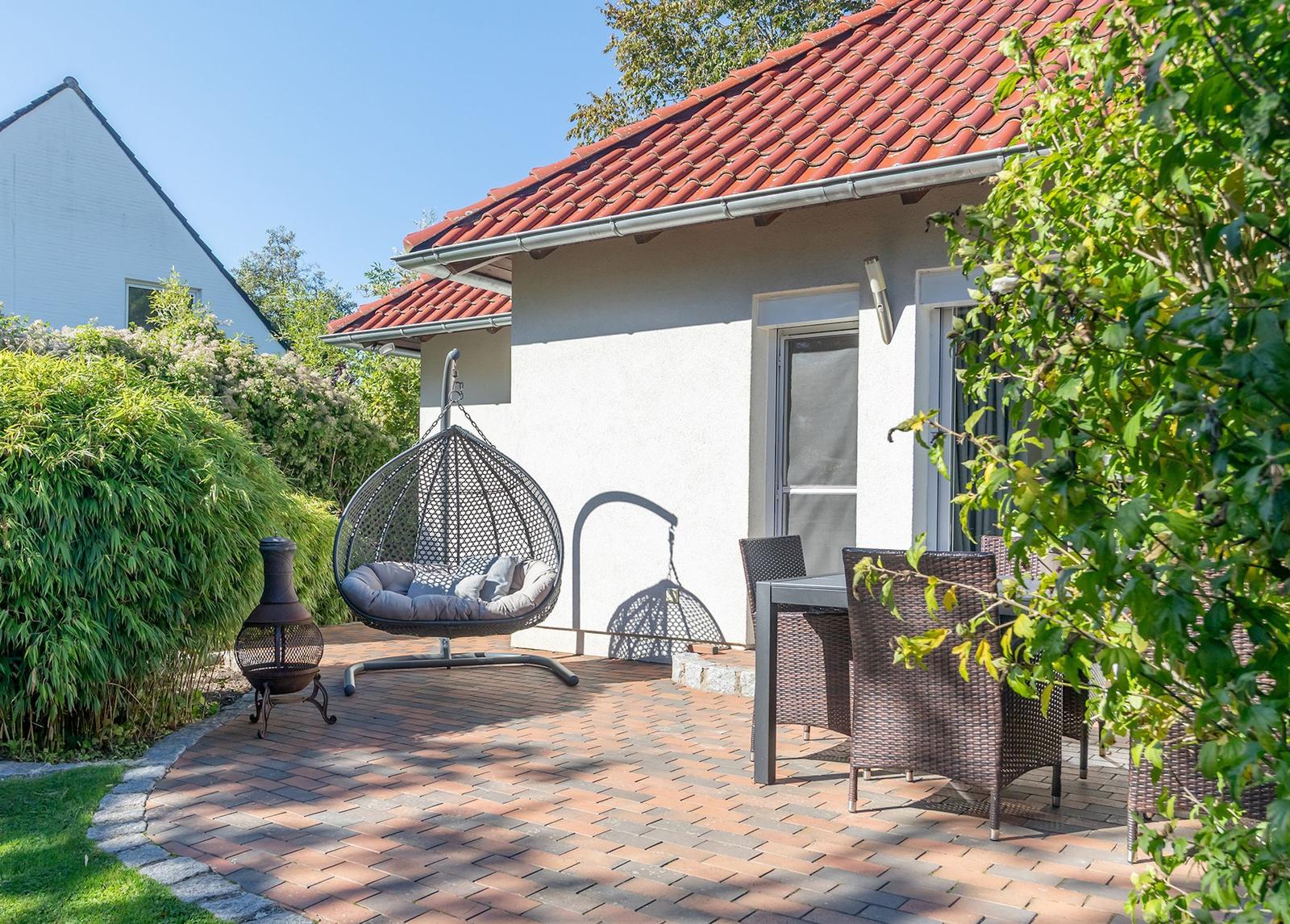 Terrasse mit Hängesessel, Tisch und Stühlen, umgeben von Grün und einem Haus mit rotem Dach.