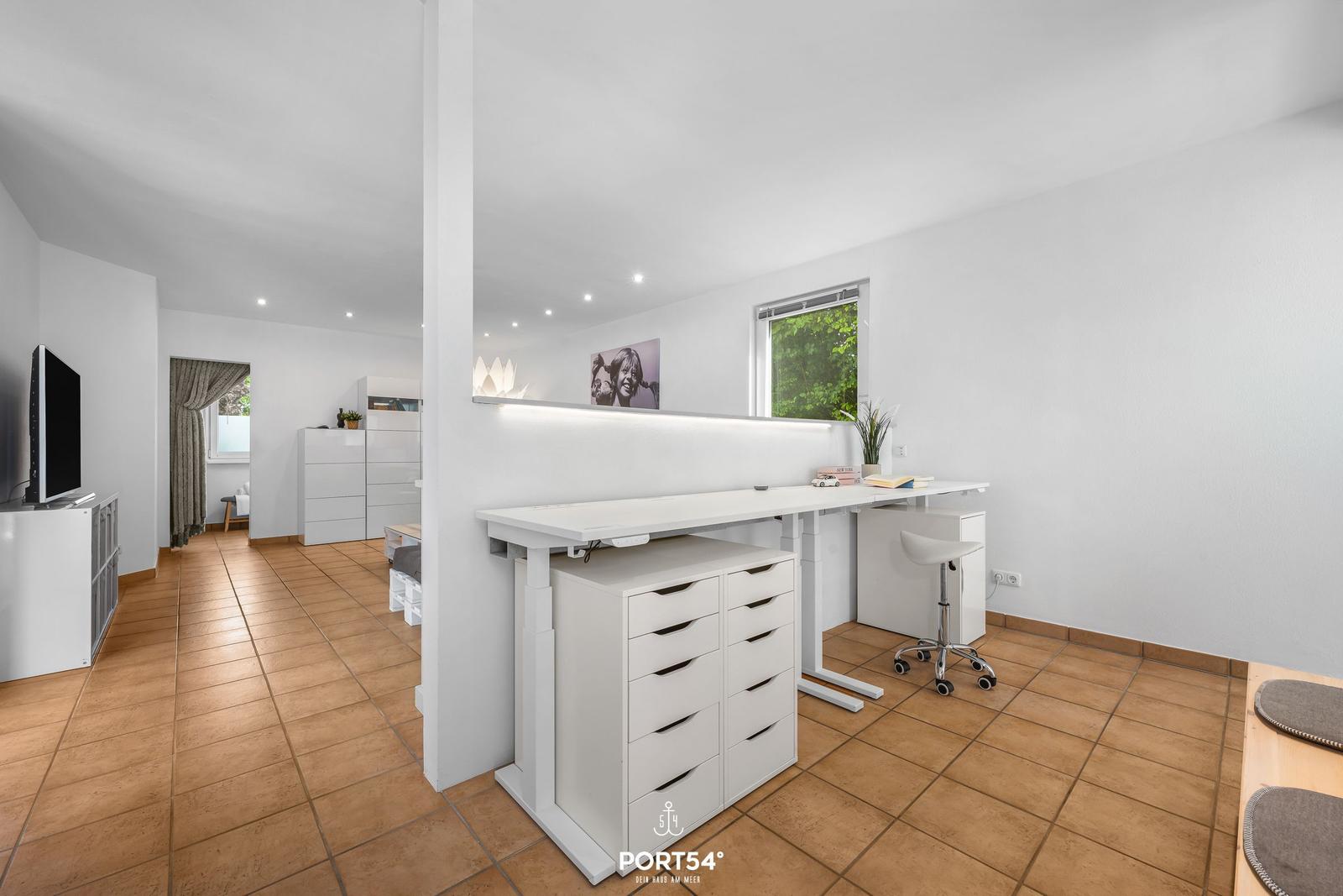 Helle Arbeitsfläche mit Stuhl und Schubladen. Im Hintergrund TV und Schlafzimmer.